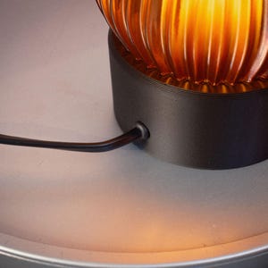 Peut inclure: Une lampe de table dot&eacute;e d'un abat-jour en verre ambr&eacute; nervur&eacute; diffusant une lueur chaude. L'abat-jour repose sur une base cylindrique noir mat. Un fin cordon d'alimentation noir sort du c&ocirc;t&eacute; de la base pour se connecter au composant &eacute;lectrique.