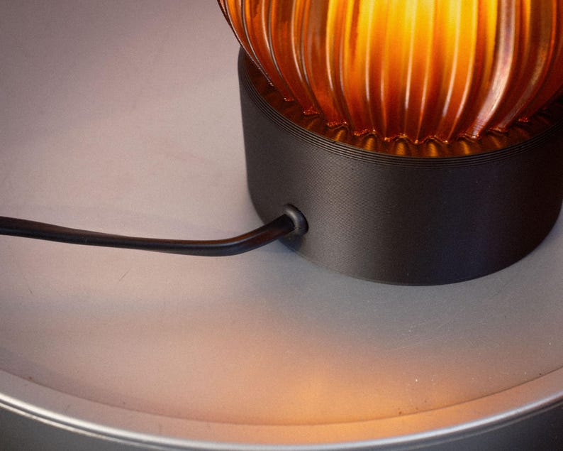 Peut inclure: Une lampe de table dot&eacute;e d'un abat-jour en verre ambr&eacute; nervur&eacute; diffusant une lueur chaude. L'abat-jour repose sur une base cylindrique noir mat. Un fin cordon d'alimentation noir sort du c&ocirc;t&eacute; de la base pour se connecter au composant &eacute;lectrique.