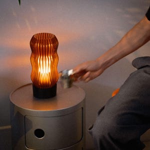Peut inclure: Une lampe de table cylindrique diffusant une lumi&egrave;re ambr&eacute;e chaleureuse &agrave; travers un abat-jour orange translucide aux nervures verticales. La lampe poss&egrave;de une base circulaire noire solide et repose sur une table d'appoint cylindrique gris clair.