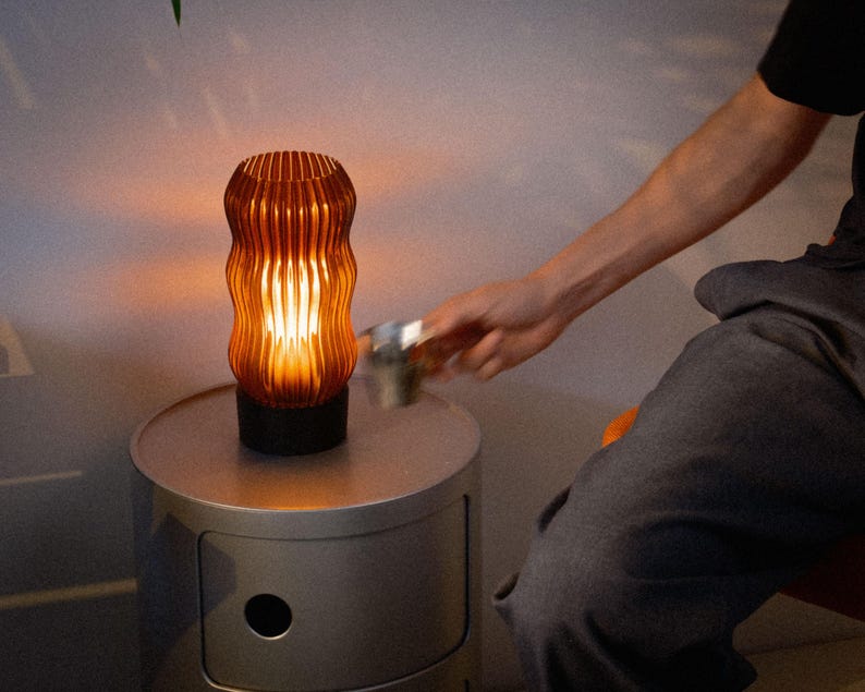 Peut inclure: Une lampe de table cylindrique diffusant une lumi&egrave;re ambr&eacute;e chaleureuse &agrave; travers un abat-jour orange translucide aux nervures verticales. La lampe poss&egrave;de une base circulaire noire solide et repose sur une table d'appoint cylindrique gris clair.