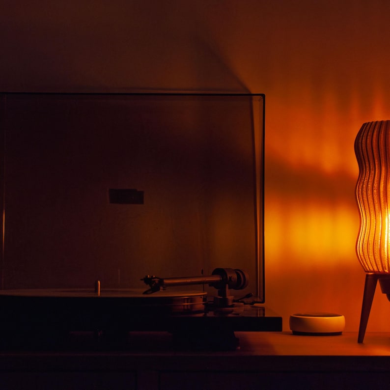 Warm ambient glow from the Wavy x Amber L lamp provides soft lighting next to a dark TV screen and turntable. Perfect mood light for media rooms. Funky retro MCM style lamp made from textured, eco-friendly recycled plastic with space-age base.