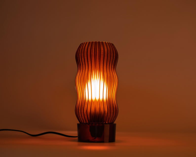 Peut inclure: Une lampe de table cylindrique avec un abat-jour translucide de couleur ambre, dot&eacute; de nervures verticales. La lampe &eacute;met une lumi&egrave;re chaude et &eacute;clatante de l'int&eacute;rieur, illuminant la surface textur&eacute;e. Elle poss&egrave;de une base sombre et un cordon noir.