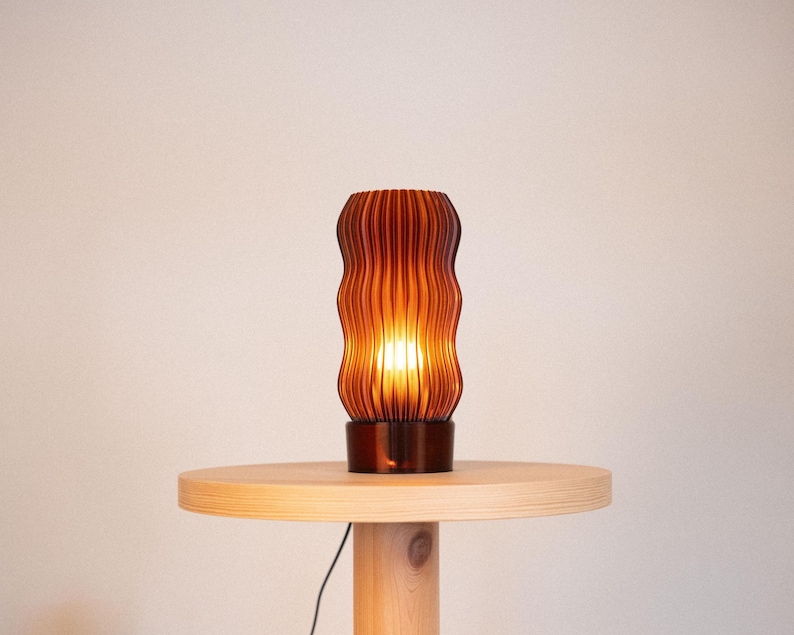 Peut inclure: Une lampe de table cylindrique avec un abat-jour translucide de couleur ambre, dot&eacute; de nervures verticales, &eacute;mettant une lumi&egrave;re chaude et &eacute;clatante. La lampe poss&egrave;de une base circulaire marron fonc&eacute; et repose sur un pi&eacute;destal en bois clair.