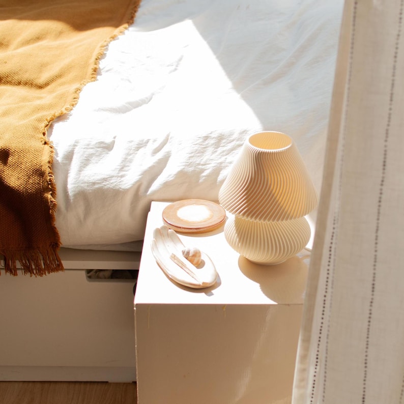 DUNE table lamp shown unlit in a bright bedroom setting on a bedside stand. Its sculptural, wavy form and soft beige color complement Organic Modern, Japandi, or even cozy minimalist decor styles. Handmade accent light made from sustainable recycled plastic with a unique texture.