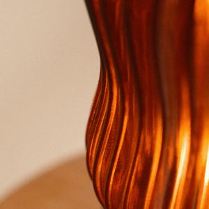 Op de afbeelding: Close-up van een getextureerd, oranjebruin object met verticale, gebogen lijnen. Het object heeft een metallic glans, die licht reflecteert en highlights en schaduwen cre&euml;ert. De achtergrond is een zachte, gebroken witte kleur.