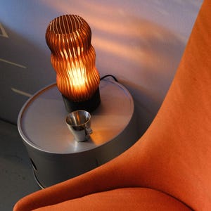 Peut inclure: Une lampe de table cylindrique avec un abat-jour nervur&eacute; de couleur ambre et une base noire, &eacute;mettant une lueur chaude. &Agrave; c&ocirc;t&eacute; de la lampe se trouve une petite tasse en m&eacute;tal argent&eacute;. L'ensemble est pos&eacute; sur une table d'appoint ronde en m&eacute;tal gris clair.