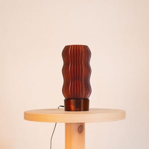 Peut inclure: Une lampe de table cylindrique pr&eacute;sentant un ext&eacute;rieur brun rouge&acirc;tre fonc&eacute; avec une s&eacute;rie de cr&ecirc;tes verticales ondul&eacute;es. La lampe poss&egrave;de une base solide et lisse ainsi qu'un fin cordon d'alimentation noir sortant de la base.