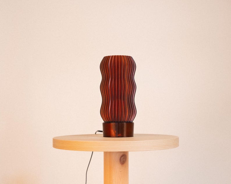 Peut inclure: Une lampe de table cylindrique pr&eacute;sentant un ext&eacute;rieur brun rouge&acirc;tre fonc&eacute; avec une s&eacute;rie de cr&ecirc;tes verticales ondul&eacute;es. La lampe poss&egrave;de une base solide et lisse ainsi qu'un fin cordon d'alimentation noir sortant de la base.