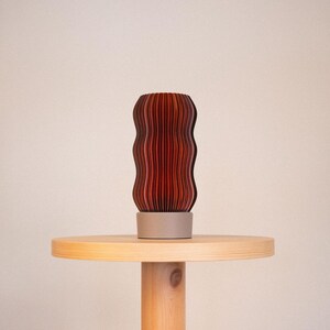 Peut inclure: Une lampe cylindrique avec un ext&eacute;rieur ondul&eacute; et nervur&eacute; verticalement, pr&eacute;sentant un d&eacute;grad&eacute; de tons rouge fonc&eacute; &agrave; brun fonc&eacute;. La lampe est soutenue par une base circulaire solide gris clair qui offre une assise stable &agrave; la structure textur&eacute;e.
