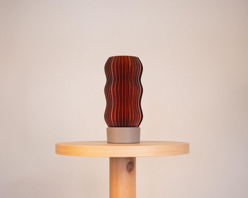 Peut inclure: Une lampe cylindrique avec un ext&eacute;rieur ondul&eacute; et nervur&eacute; verticalement, pr&eacute;sentant un d&eacute;grad&eacute; de tons rouge fonc&eacute; &agrave; brun fonc&eacute;. La lampe est soutenue par une base circulaire solide gris clair qui offre une assise stable &agrave; la structure textur&eacute;e.