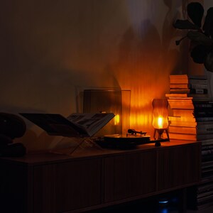 Wavy x Amber L lamp glows warmly, made from amber recycled plastic. Shown lit on a sideboard beside a vinyl record player & books, creating perfect mood lighting. Ideal retro, mid-century modern, or funky accent for cozy spaces. Eco-friendly choice.