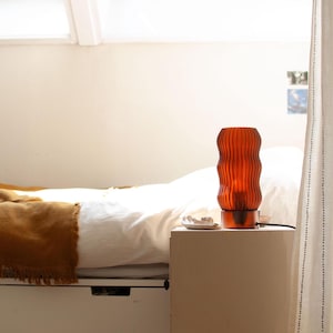 Handmade WAVY x AMBER accent lamp glowing on a bedroom bedside table. Features unique 3D printed fluted amber shade providing warm ambient light. Perfect table lamp for cozy home decor and mood lighting. Modern design by 3Deco Studio shown in naturally lit room setting next to bed.
