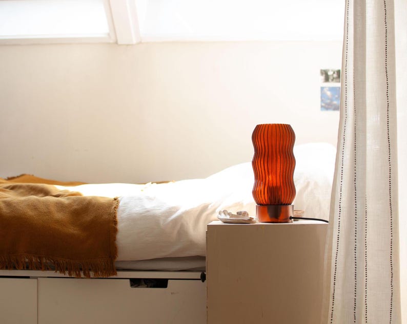Handmade WAVY x AMBER accent lamp glowing on a bedroom bedside table. Features unique 3D printed fluted amber shade providing warm ambient light. Perfect table lamp for cozy home decor and mood lighting. Modern design by 3Deco Studio shown in naturally lit room setting next to bed.