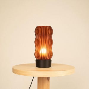 Peut inclure: Une lampe de table cylindrique dot&eacute;e d'un abat-jour brun fonc&eacute; &agrave; la texture ondul&eacute;e et aux nervures verticales, diffusant une lumi&egrave;re chaude et &eacute;clatante. L'abat-jour est fix&eacute; sur une base noire circulaire solide, cr&eacute;ant un contraste marqu&eacute;.