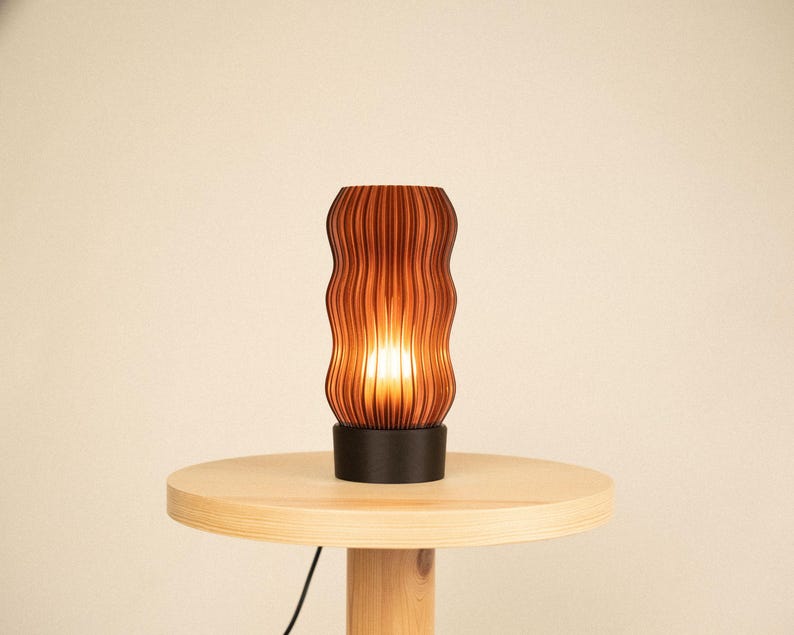 Peut inclure: Une lampe de table cylindrique dot&eacute;e d'un abat-jour brun fonc&eacute; &agrave; la texture ondul&eacute;e et aux nervures verticales, diffusant une lumi&egrave;re chaude et &eacute;clatante. L'abat-jour est fix&eacute; sur une base noire circulaire solide, cr&eacute;ant un contraste marqu&eacute;.