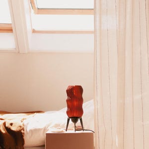 Retro Wavy x Amber L table lamp sits bedside in a bright room. Features unique wavy texture in warm amber recycled plastic and a sleek black tripod base. Mid-century modern meets space age design for cozy, eco-conscious mood lighting.