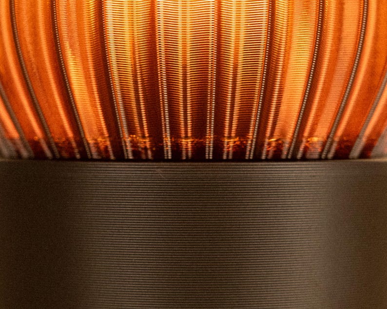 Peut inclure: Une vue rapproch&eacute;e d'une lampe avec un abat-jour en verre ambr&eacute; translucide dot&eacute; de textures nervur&eacute;es verticales. L'abat-jour repose sur une base cylindrique noire mate solide qui pr&eacute;sente de fines lignes horizontales sur sa surface.