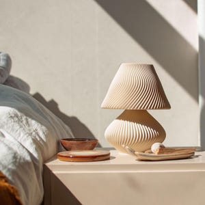 Lifestyle photo of the Japandi-style mushroom lamp used as a piece of sculptural decor on a modern bedside table. Shown unlit in bright, natural morning light, the lamp's unique wavy texture interacts with the strong shadows from a nearby window. This demonstrates its appeal as a stylish object in a serene, Scandinavian-inspired bedroom.