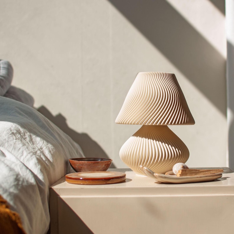 Lifestyle photo of the Japandi-style mushroom lamp used as a piece of sculptural decor on a modern bedside table. Shown unlit in bright, natural morning light, the lamp's unique wavy texture interacts with the strong shadows from a nearby window. This demonstrates its appeal as a stylish object in a serene, Scandinavian-inspired bedroom.