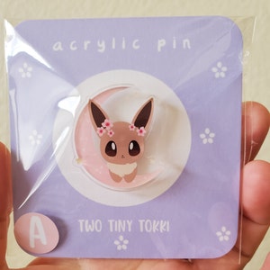 May include: A purple card with the text "acrylic pin" and "TWO TINY TOKKI" on it. A clear plastic circle with a cartoon image of a brown Eevee with pink flowers on its head, sitting on a crescent moon, is on the card. The letter "A" is in a circle on the bottom left corner of the card.
