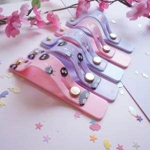 May include: A selection of pastel-coloured phone grips with cartoon character designs. Each grip features a different colour scheme, including pink, purple, and blue, with gold-coloured button accents. The grips are arranged on a light pink surface.