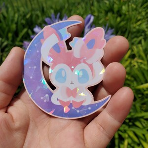 Puede incluir: Una pegatina hologr&aacute;fica con una criatura de dibujos animados con detalles rosas, blancos y azules, sentada en una luna creciente. La luna tiene un degradado p&uacute;rpura y azul con constelaciones.