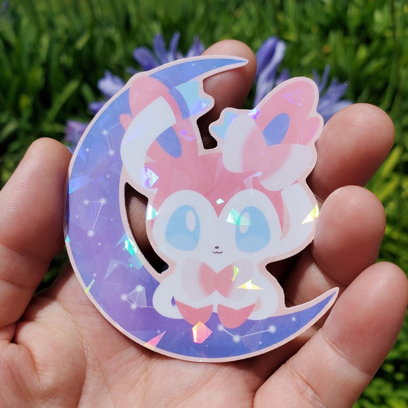 Puede incluir: Una pegatina hologr&aacute;fica con un personaje de dibujos animados en rosa, blanco y azul, sentado en una luna creciente. La luna es morada con un dise&ntilde;o de constelaci&oacute;n. La pegatina tiene un acabado brillante e iridiscente.