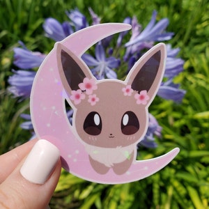 May include: A sticker featuring a cartoon character with large eyes, sitting on a pink crescent moon. The character has brown fur, long ears, and a floral crown. The moon has a constellation design.