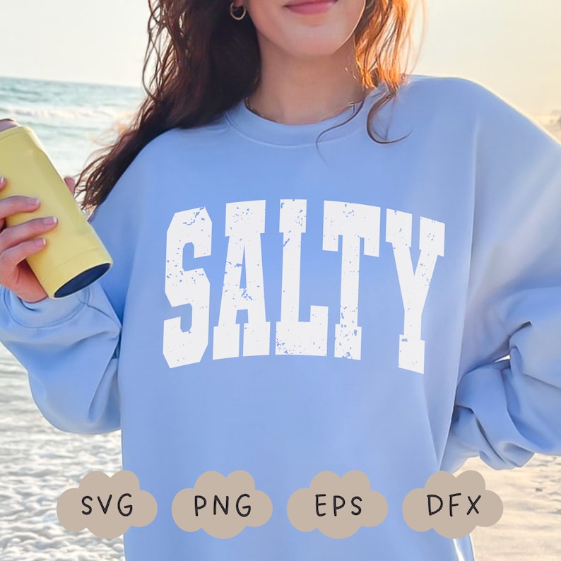 May include: A light blue sweatshirt with the word "SALTY" printed in white with a distressed, vintage look. The sweatshirt is worn by a person standing in front of a beach scene.