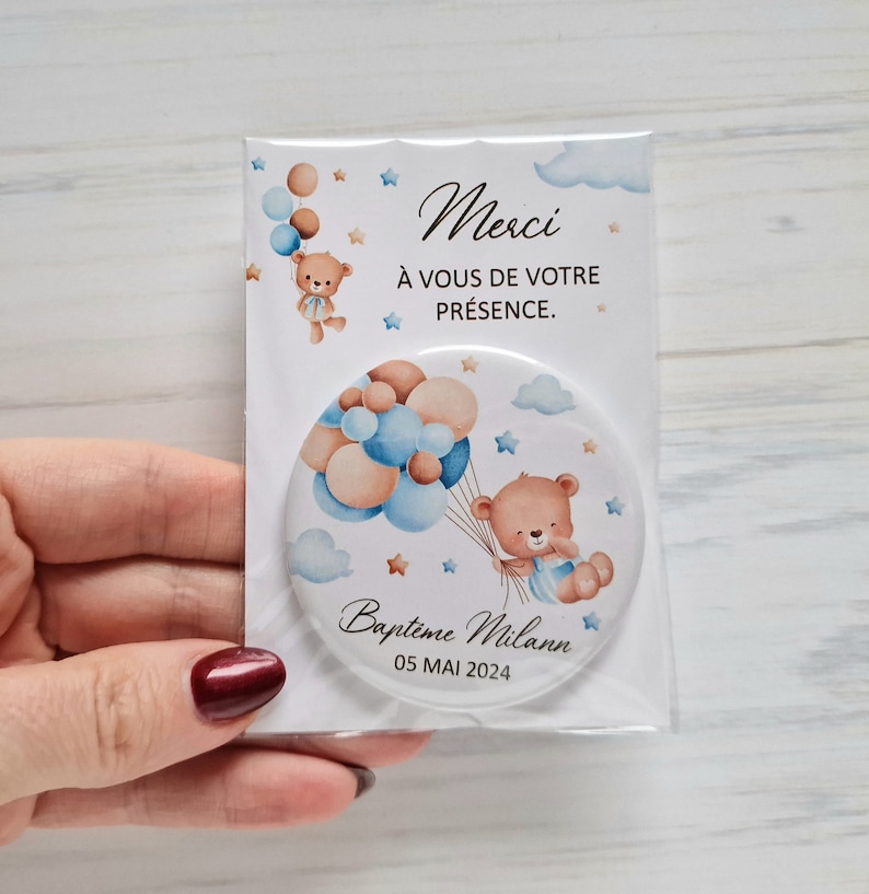 May include: A round mirror with a white background and a teddy bear holding blue and brown balloons. The mirror has the text "Baptime Milan 05 MAI 2024" and "Merci &agrave; vous de votre pr&eacute;sence."