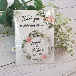 May include: A circular pin-back button featuring a gold geometric frame, pink roses, and green leaves. The text reads Amanda & Frederick and April 23, 2022. It is packaged on a card that says Thank you, for celebrating with us! in black script.