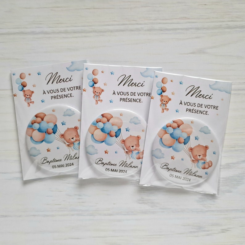 May include: Three round blue and brown favor buttons with a teddy bear holding balloons. The buttons have the text "Merci" and "A vous de votre presence" and "Bapteme Milann 05 MAI 2024".