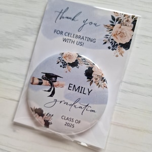 Könnte beinhalten: Ein runder, blau-weißer Party-Favor für den Abschluss mit floralem Design. Der Favorit zeigt eine schwarze Abschluss-Kappe und eine Schriftrolle, den Namen "Emily", das Wort "Abschluss" und den Text "Klasse von 2025".