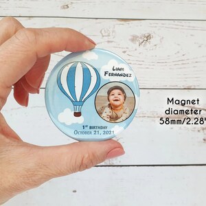 May include: A blue and white hot air balloon with clouds and a photo of a baby boy on a round magnet. The magnet says "Liam Fernandez" and "1st Birthday October 21, 2021". The magnet is 2.28 inches in diameter.