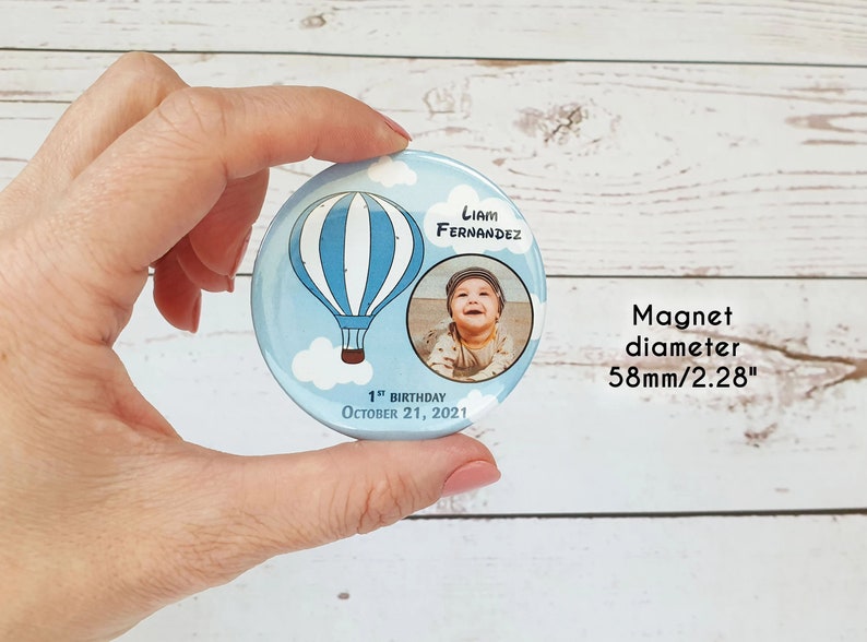 May include: A blue and white hot air balloon with clouds and a photo of a baby boy on a round magnet. The magnet says "Liam Fernandez" and "1st Birthday October 21, 2021". The magnet is 2.28 inches in diameter.