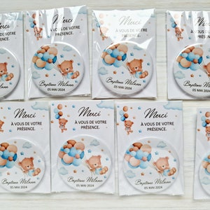 May include: A set of round, white,  magnet-style favors with a blue and brown teddy bear holding balloons. The text on the favors reads "Merci &agrave; vous de votre pr&eacute;sence. Baptime Milann 05 MAI 2024".