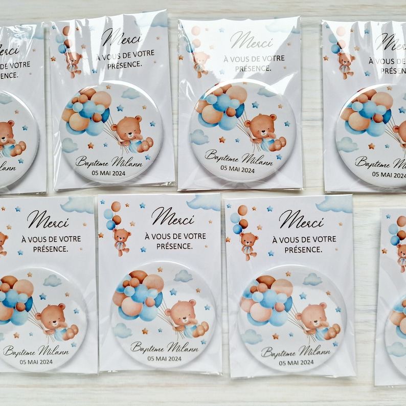 May include: A set of round, white,  magnet-style favors with a blue and brown teddy bear holding balloons. The text on the favors reads "Merci &agrave; vous de votre pr&eacute;sence. Baptime Milann 05 MAI 2024".