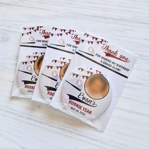 Könnte beinhalten: Drei Baseball-Partygeschenke in Klarsichtverpackung. Jedes Geschenk hat einen runden Knopf mit Baseball-Design und dem Text "Evan's Rookie Year July 19, 2025". Die Verpackung enthält die Worte "Thank you" und "For making my birthday a grand slam!"