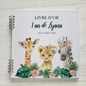 Könnte beinhalten: Weißes Spiralbuch mit Aquarellillustrationen von Giraffe, Löwenjungem und Zebra, jeweils mit Blumenkränzen. Der Text lautet "LIVRE D'OR 1 an de Lyana LE 24 AOÛT 2025" in Dunkelgrün. Tropische Blätter und Blumen umrahmen die Tiere.