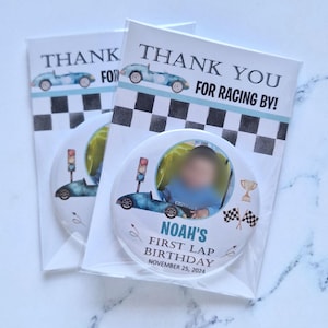 May include: A round, white button with a blue race car, a checkered flag, and a trophy. The button also has a photo of a baby in a blue shirt and the text "Noah's First Lap Birthday November 25, 2024".