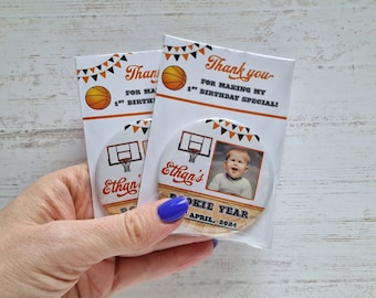 May include: Two round thank you favor tags with a basketball theme. The tags feature a basketball hoop with a photo of a baby boy in a white shirt. The text on the tags reads "Thank you for making my 1st birthday special!" and "Ethan's Rookie Year April, 2024".