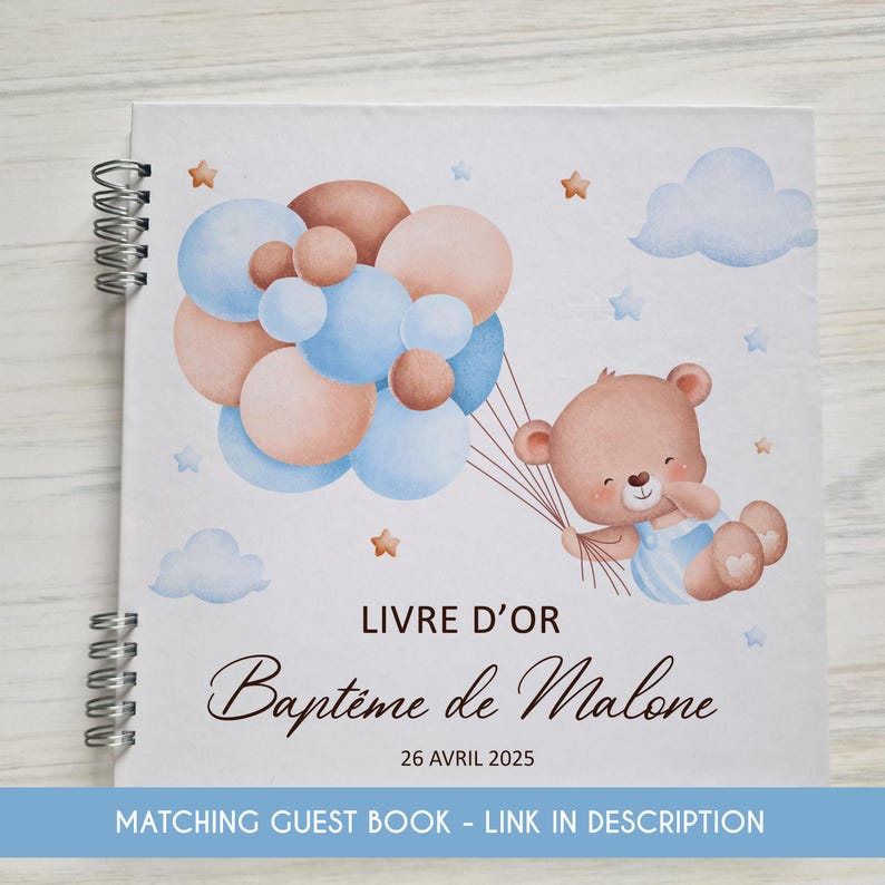 May include: A white guest book with a brown teddy bear holding blue and pink balloons. The book has the text "LIVRE D'OR Bapt&ecirc;me de Malone 26 AVRIL 2025" on the front cover.