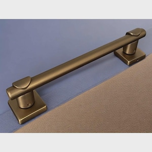May include: A bronze-colored towel bar with a rectangular base and a curved top. The bar is mounted on a blue and tan surface. The design is simple and modern, suitable for a bathroom or kitchen.