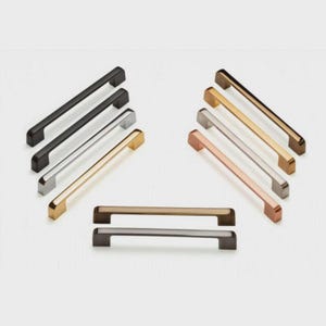 May include: A collection of cabinet pulls in various metallic finishes, including black, silver, gold, and copper. The pulls are rectangular with a bar handle design, suitable for drawers and cabinet doors. The hardware is displayed against a white background.