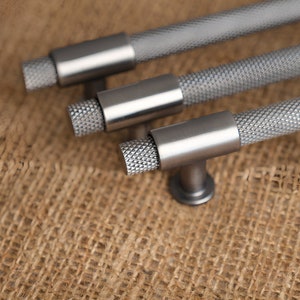 May include: Three silver cabinet pulls with a textured, diamond-patterned grip and cylindrical center. The pulls have a T-bar design and are set against a neutral, textured background. The hardware is likely for furniture or cabinetry.