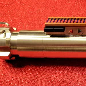 May include: A metallic lightsaber prop with silver, black, and gold accents. The hilt features a ribbed design, a rectangular control box with purple stripes, and a ring at the end. The prop is set against a red background.
