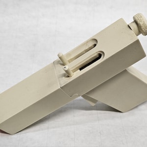 May include: A beige wooden tool with a rectangular body and a knob on one end. The tool has two small pegs and a sliding mechanism. The tool is designed for woodworking or crafts.
