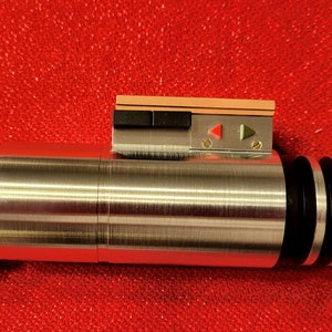 May include: A metallic silver lightsaber prop with black and red accents. The prop features a detailed handle with a ring and a control panel with buttons and indicator lights. The lightsaber is set against a red background.
