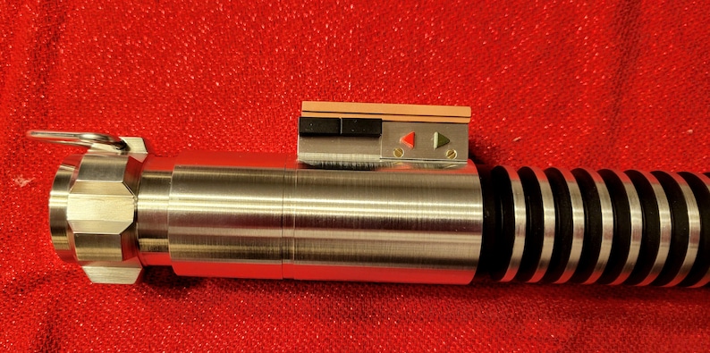 May include: A metallic silver lightsaber prop with black and red accents. The prop features a detailed handle with a ring and a control panel with buttons and indicator lights. The lightsaber is set against a red background.