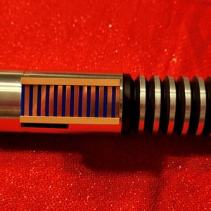 May include: A detailed, metallic lightsaber prop with a silver handle, black and silver ribbed grip, and a section with blue and copper-colored details. The prop is set against a red background.